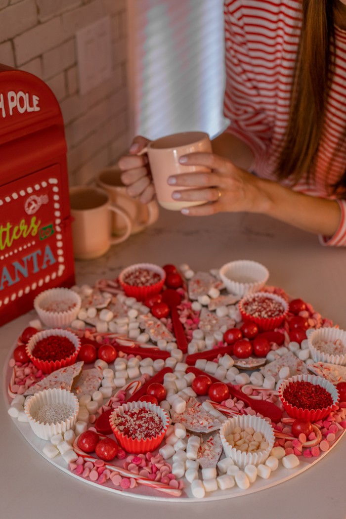 Christmas hot chocolate board with woman in background holding a pink mug. 